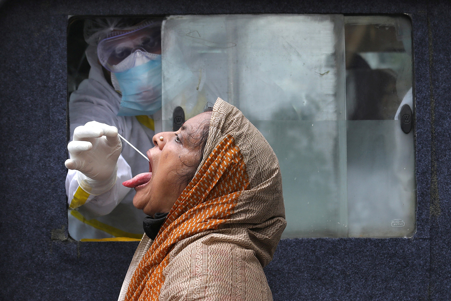 This is a striking photo showing a health worker in full protective gear reaching through a window to swab the tongue of a woman.