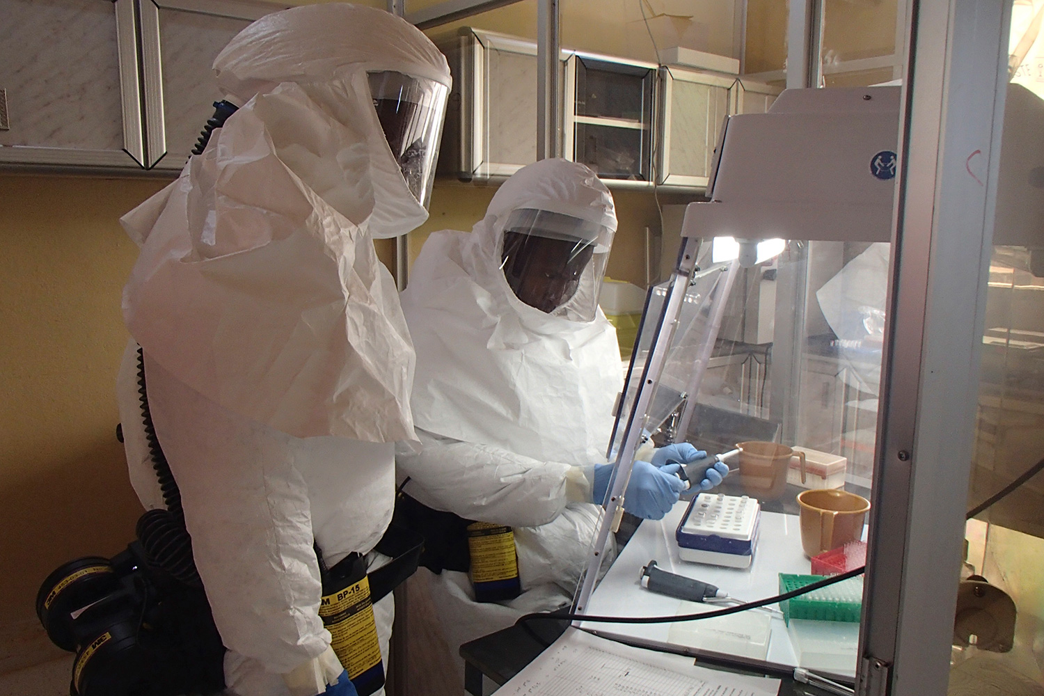The photo shows two workers in full body protective gear working under a hood.