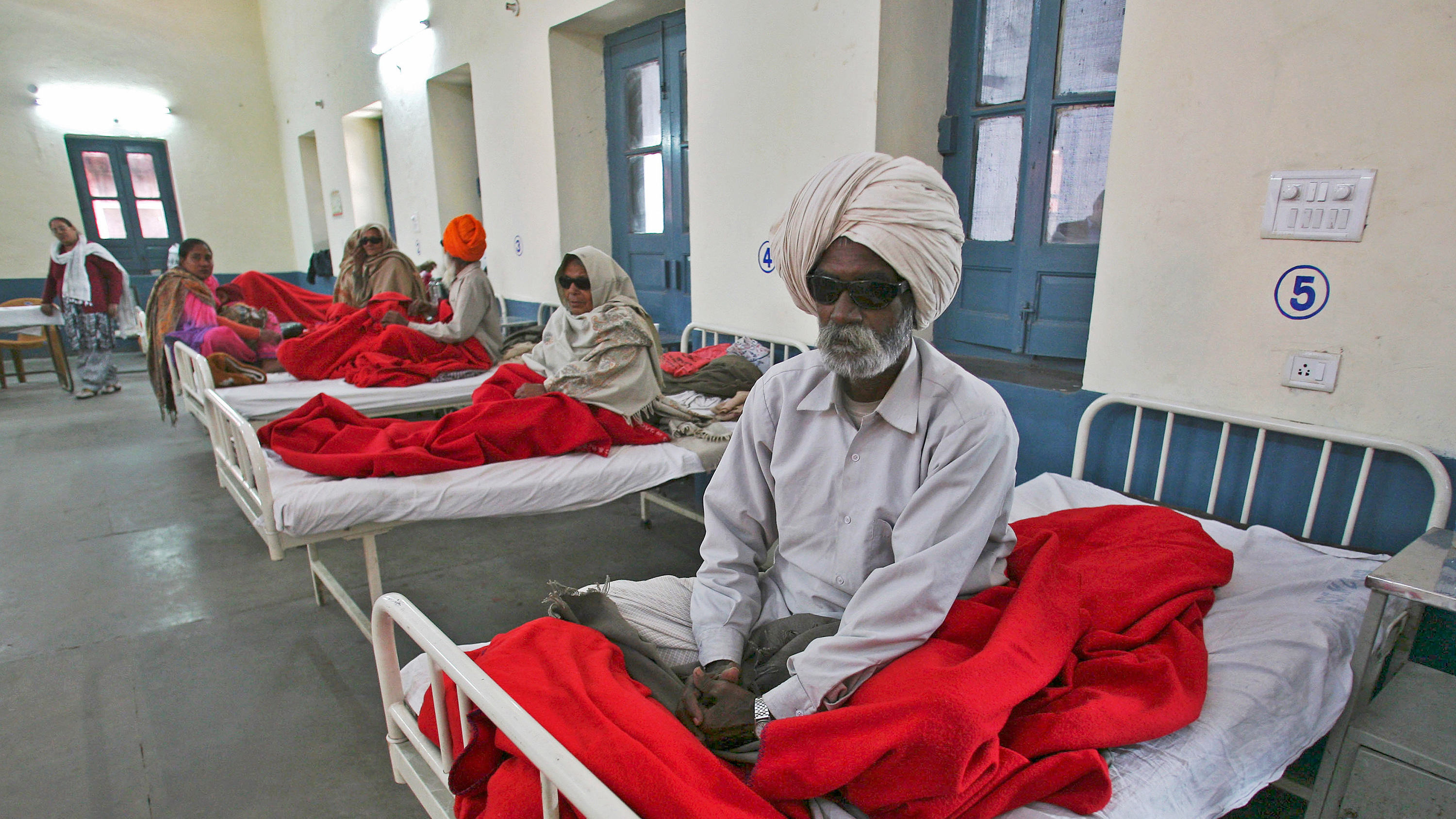 The photo shows several people sitting on hospital beds. One man in the foreground is wearing dark glasses.