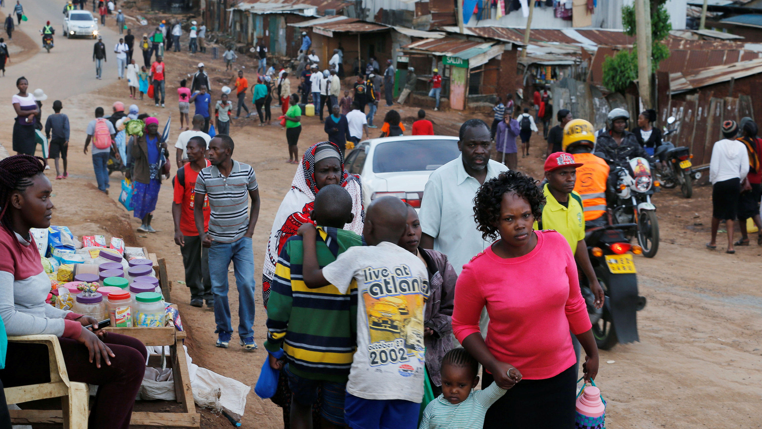 The image shows a long dirt road lined with tin shacks stretching off in the distance. Many people are walking on the road toward the camera and a woman has a table selling goods in the foreground.
