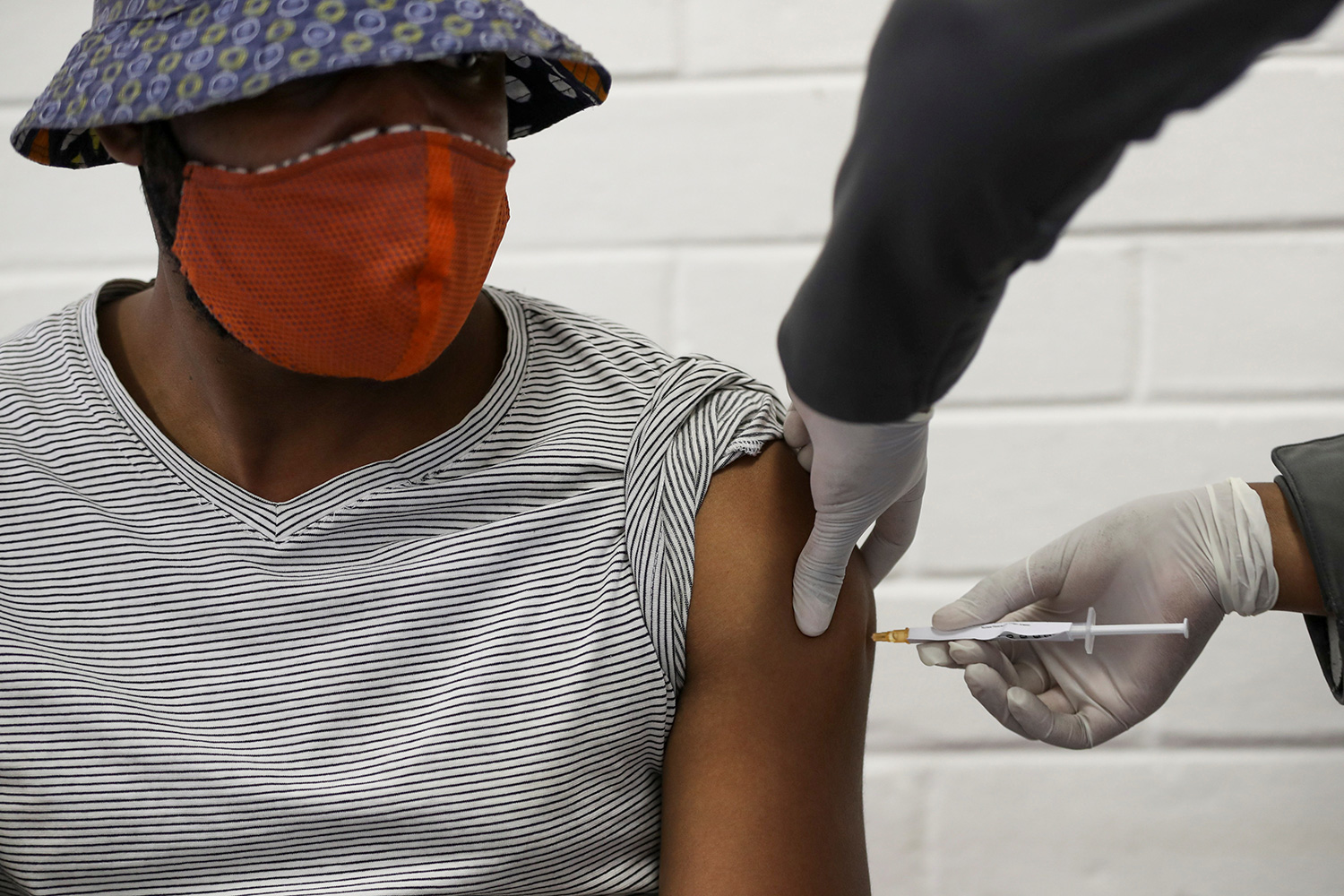 The photo shows a man with a hat and a mask getting a jab in his arm.