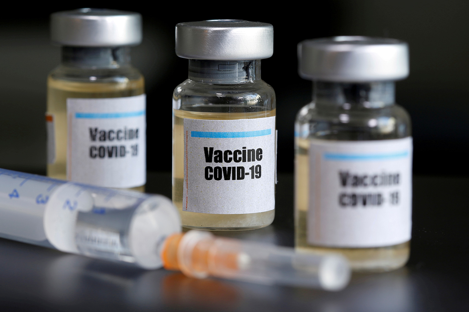 The photo shows three small vials labelled "COVID-19 Vaccine" and a syringe.