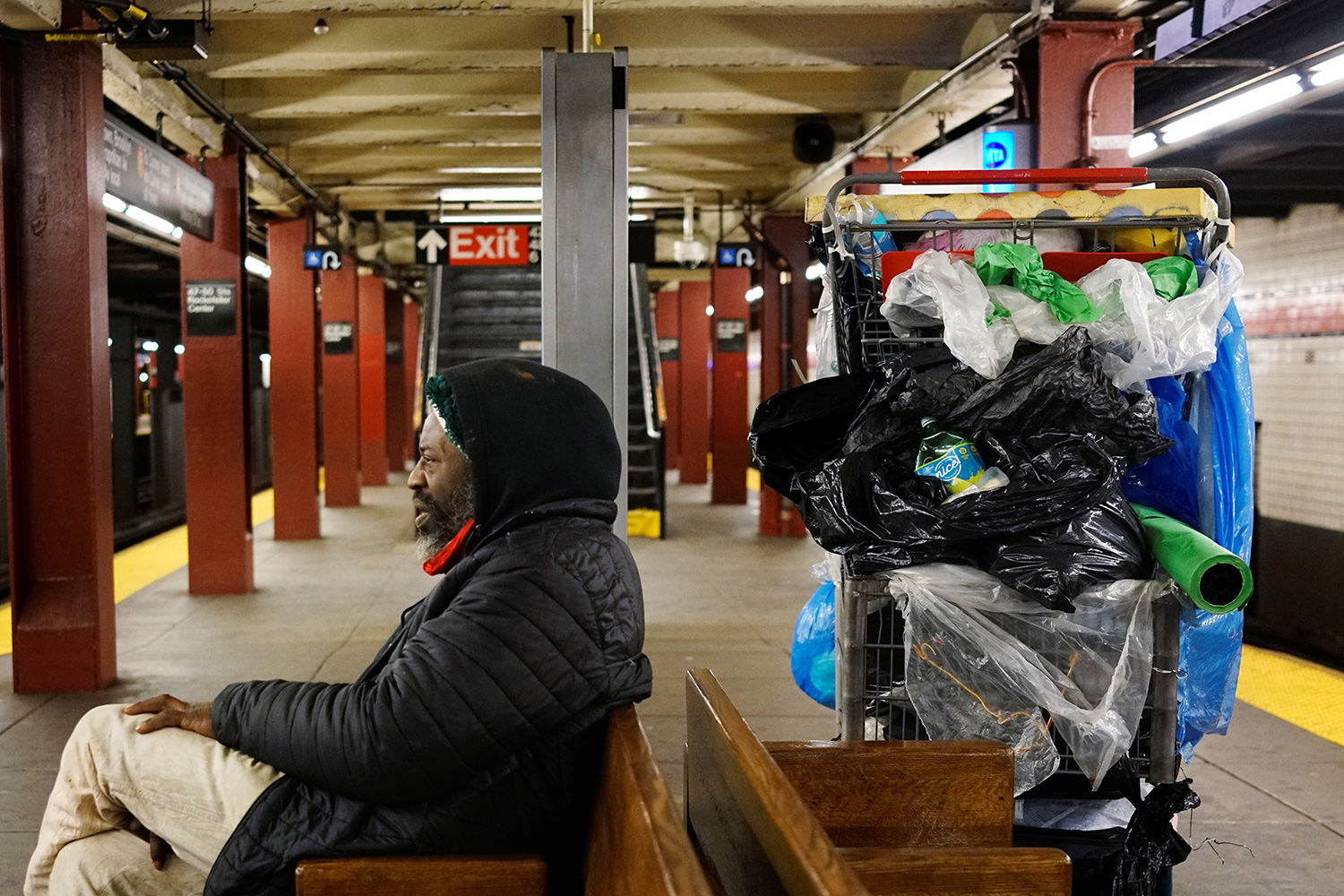 Picture shows a man sitting on a bench with large cart of stuff sitting behind him.