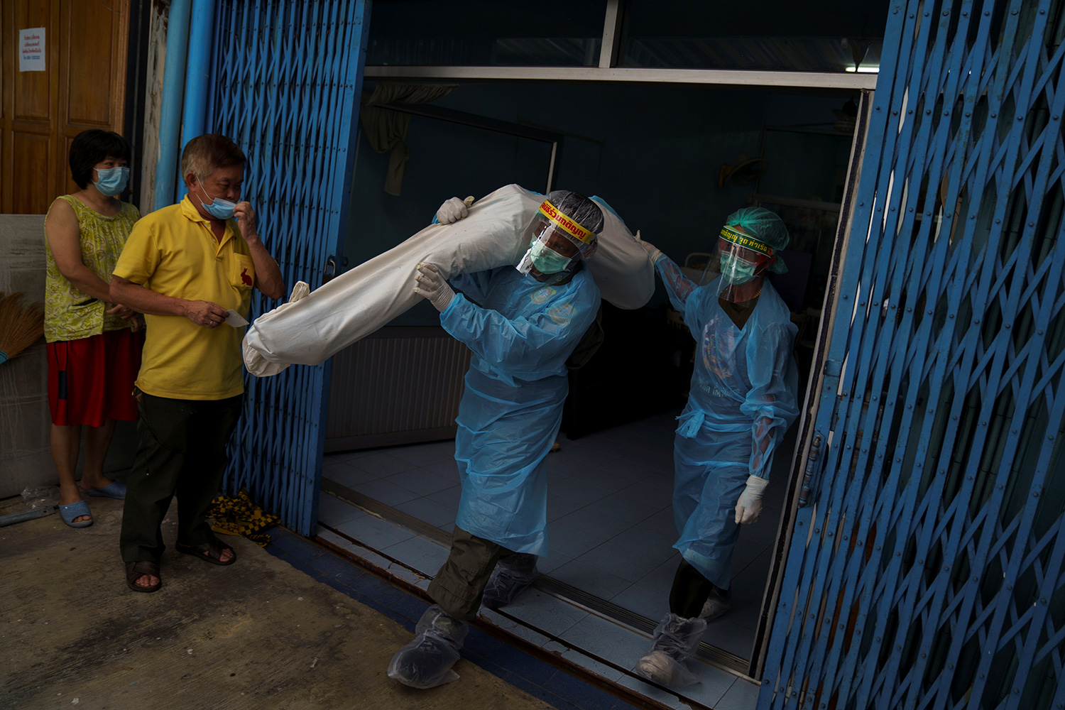The photo shows two workers in blue protective gear walking out of a building. One of them has a body wrapped in a white covering over his shoulder.