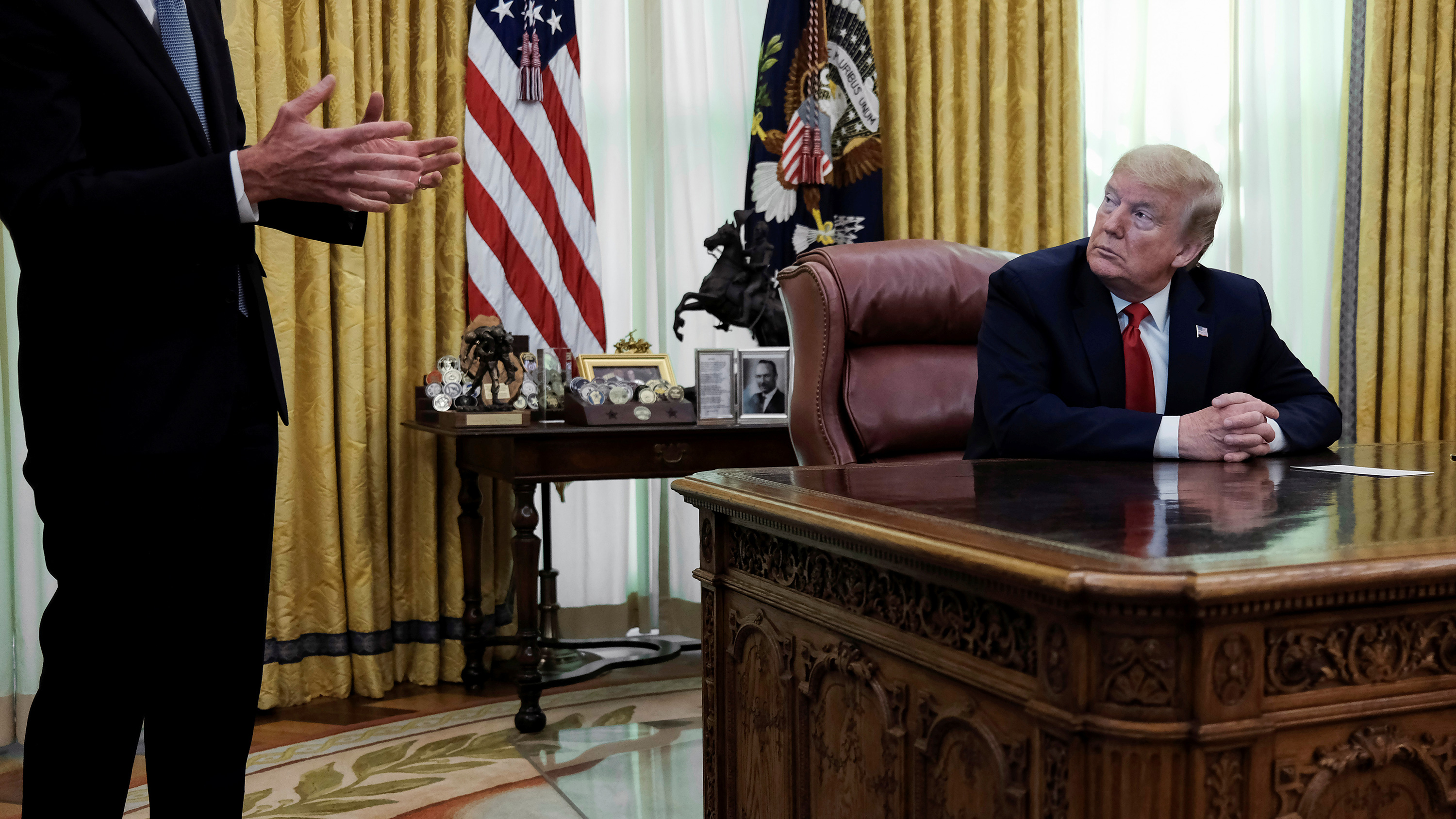 The photo shows the president at his desk looking to the right as an suited man speaks.