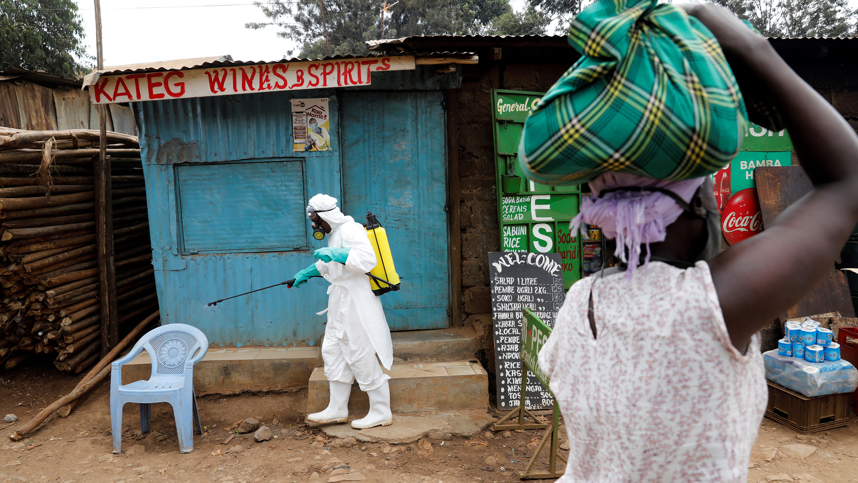 The photo shows a health worker in protective gear spraying from a bottle while a woman in the foreground looks on.