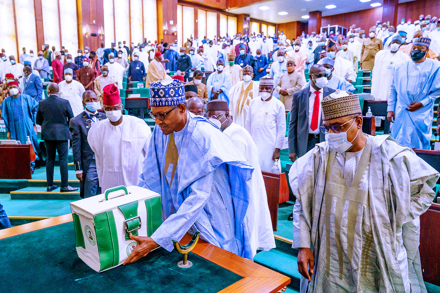 The photo shows the president placing a smart white-and-green portfolio case on a table at the front of a large assembly hall filled with people.