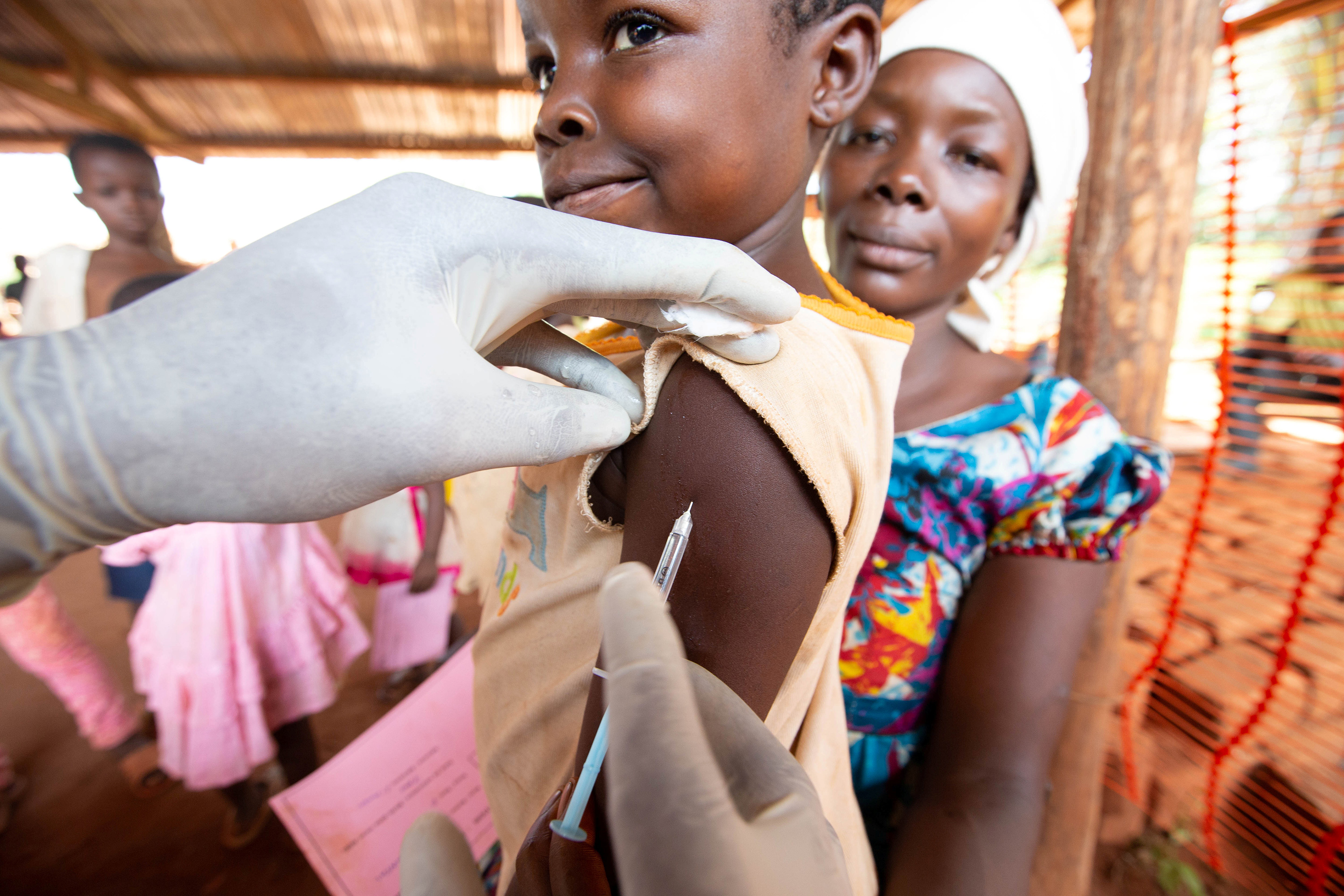 Picture shows a small child receiving a jab in the arm while an adult, presumably the child's caregiver, looks on in the background.