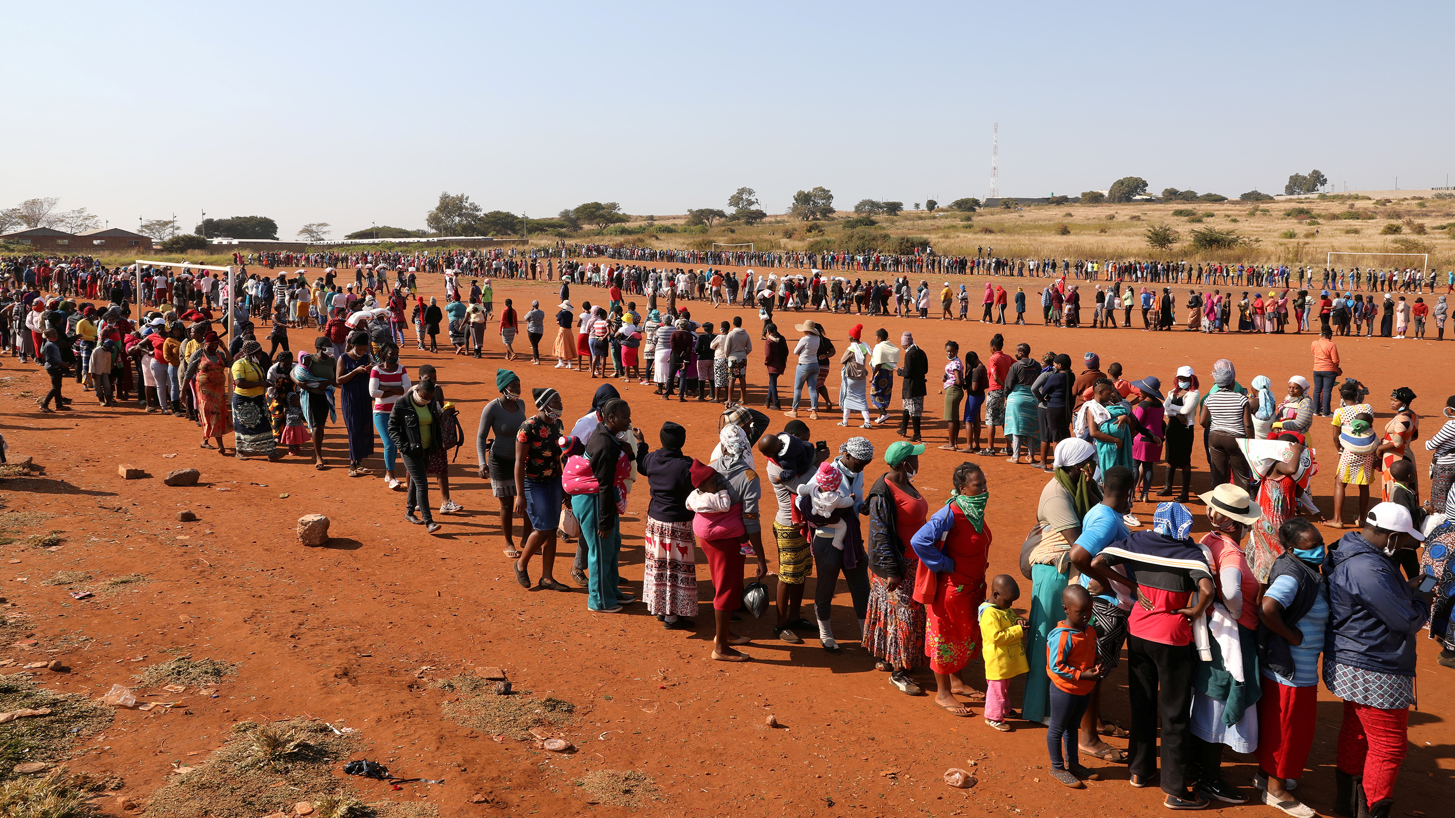 The photo is taken from a high vantage overlooking a huge crowd of people standing on line in a red soil dry plain.