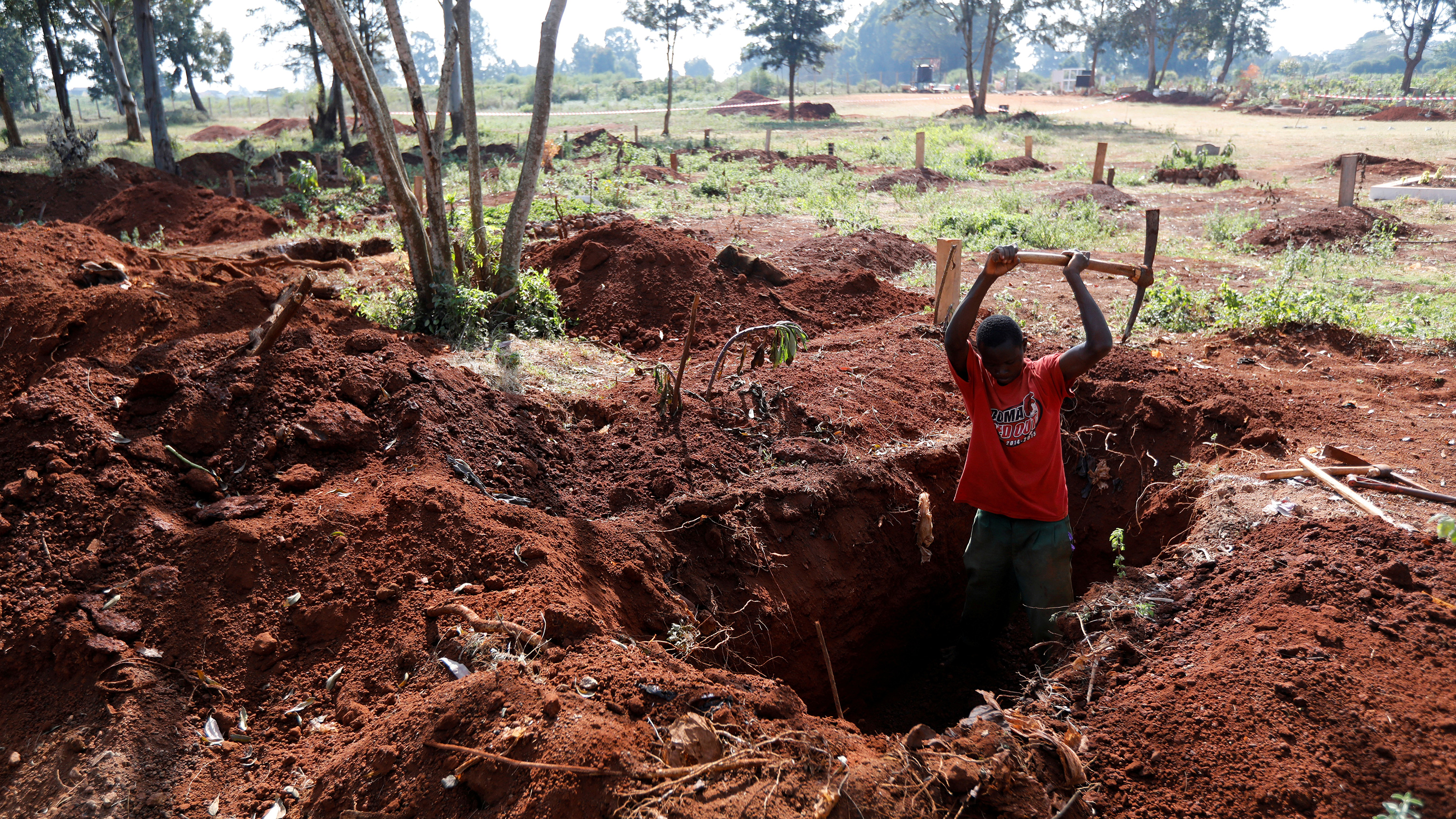 The photo shows a worker chest deep in an oblong hole actively swinging his pick into the red earth with lots of other freshly dug graves visible in the background.