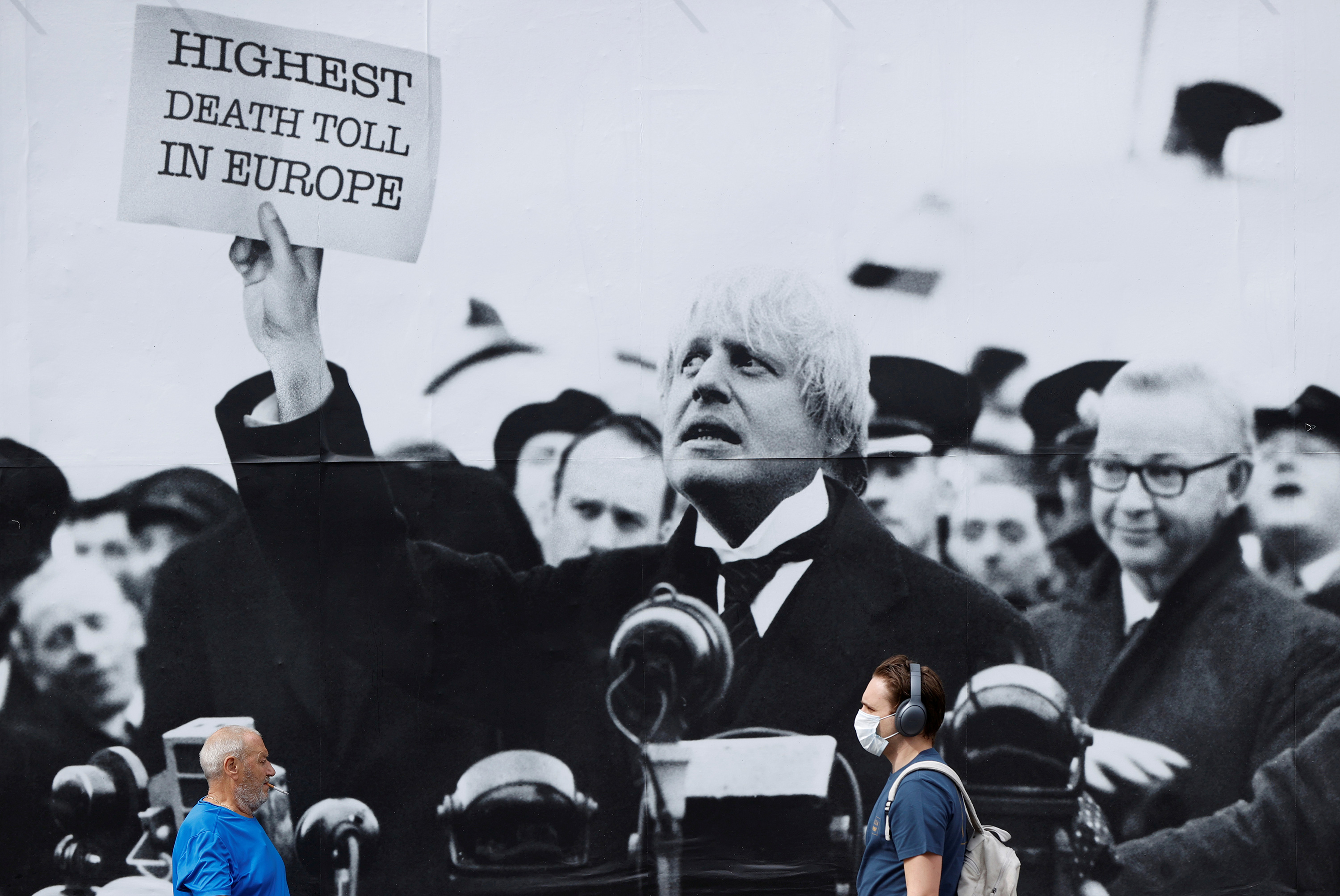 The photo shows a few people walking by a huge sign with a black and white picture of the PM holding up a placard that reads, "Highest Death Toll in Europe."