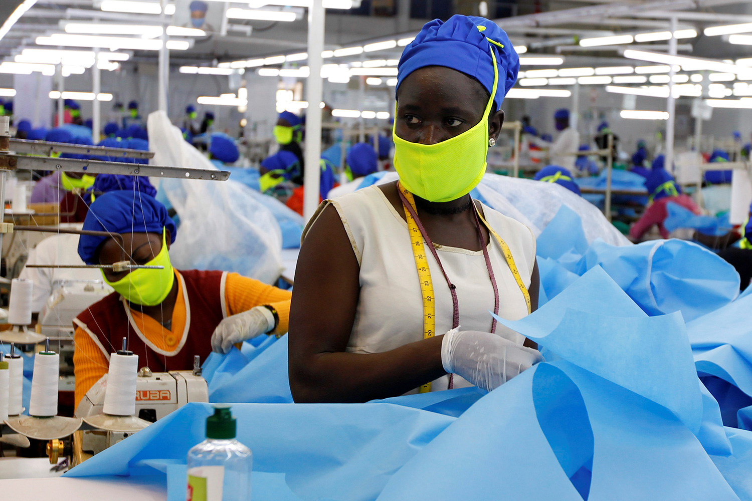 The photo shows a row of workers wearing protective blue suits and bright green facemasks working in a factory.