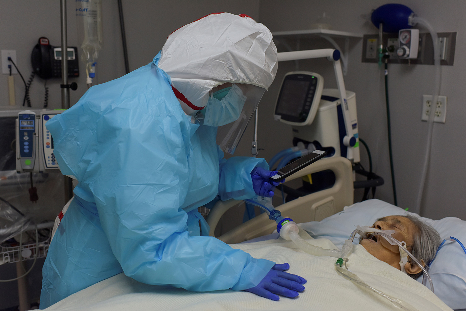 Picture shows a woman dressed in protective gear leaning over an older woman in a hospital bed with her phone in hand.