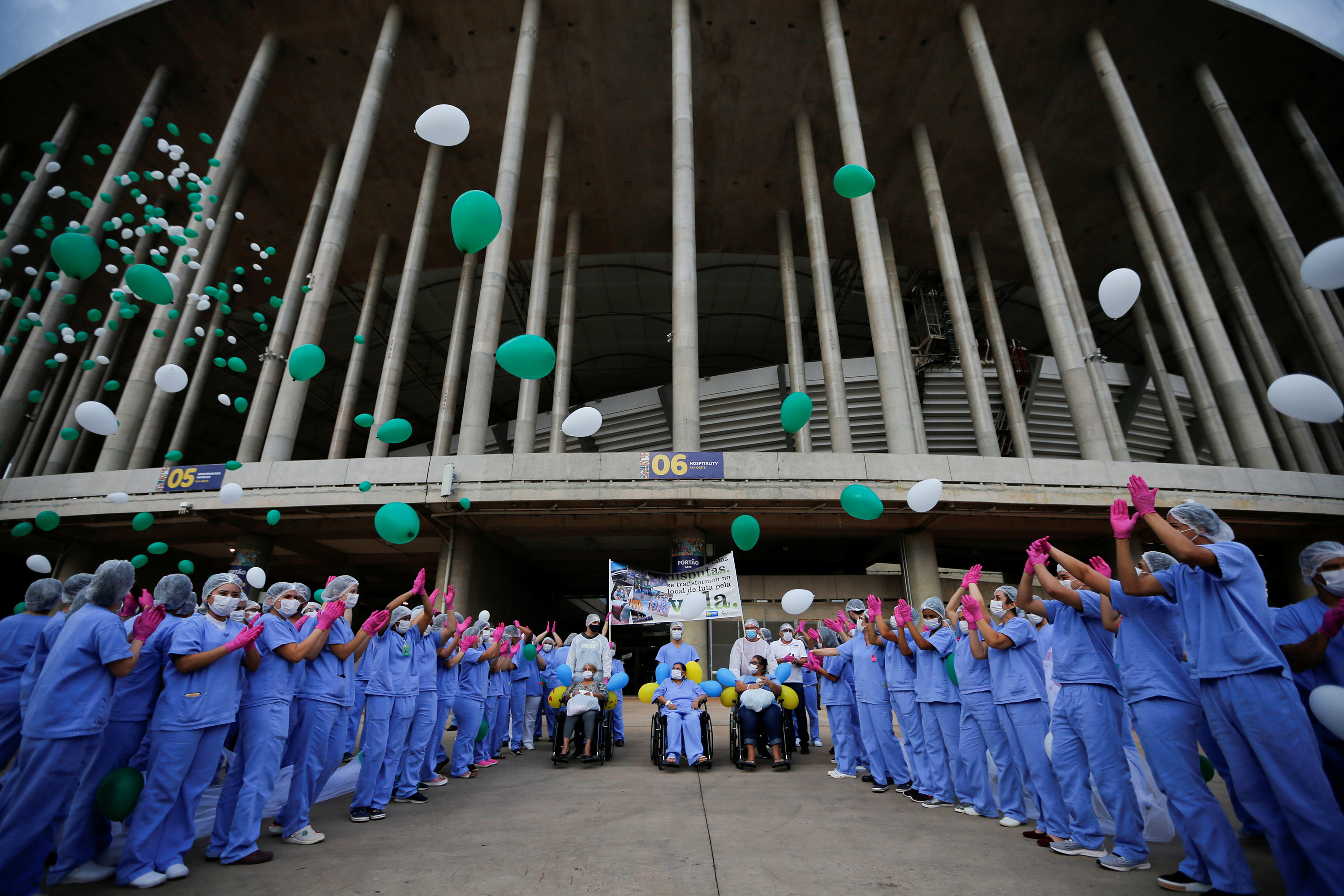 The photo shows a large crowd of blue scrubs wearing health workers cheering and releasing green and white balloons at the entrance of a large stadium.