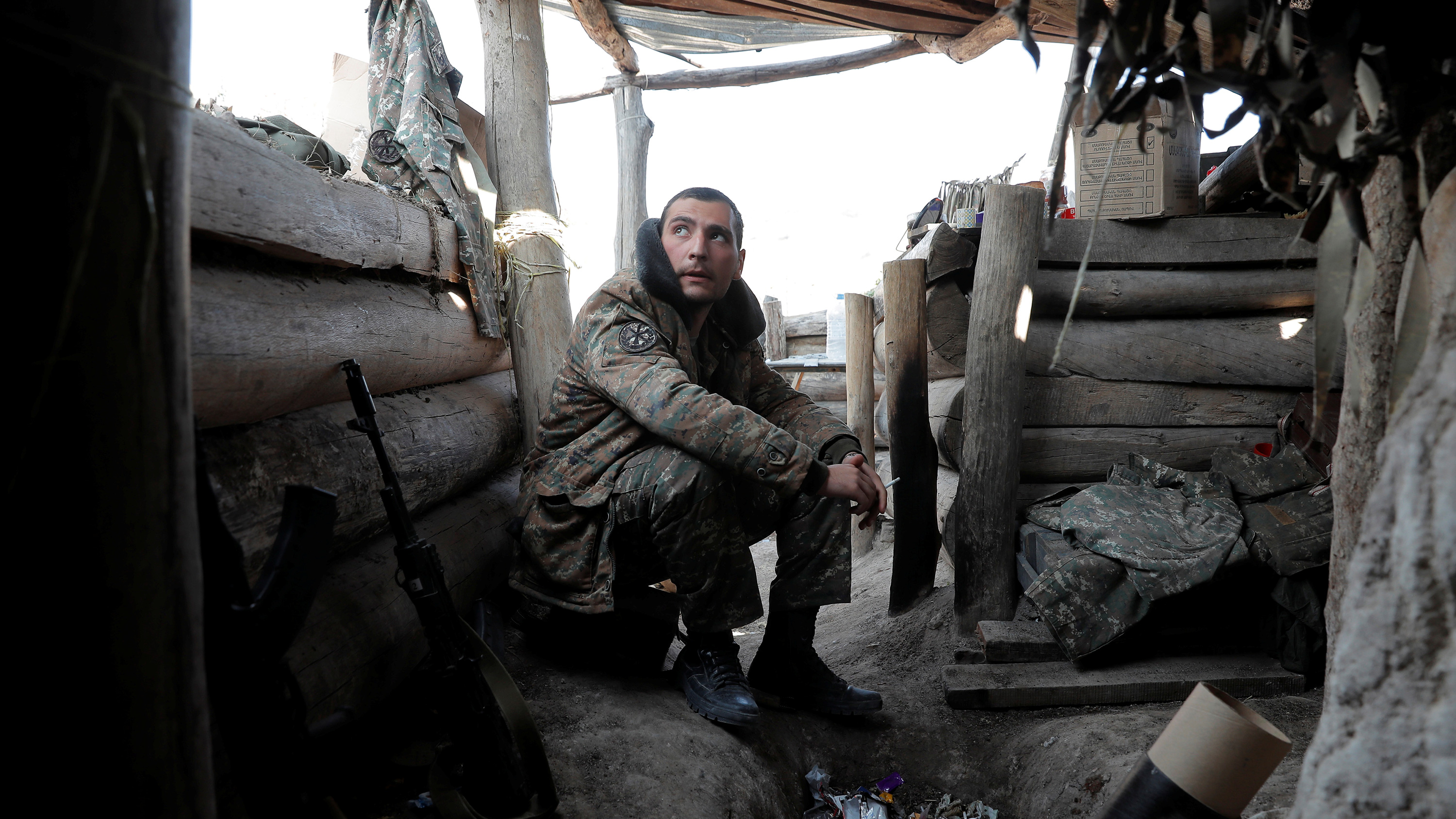 The photo shows a man crouched down in a trench smoking a cigarette and looking nervously over the battlements.