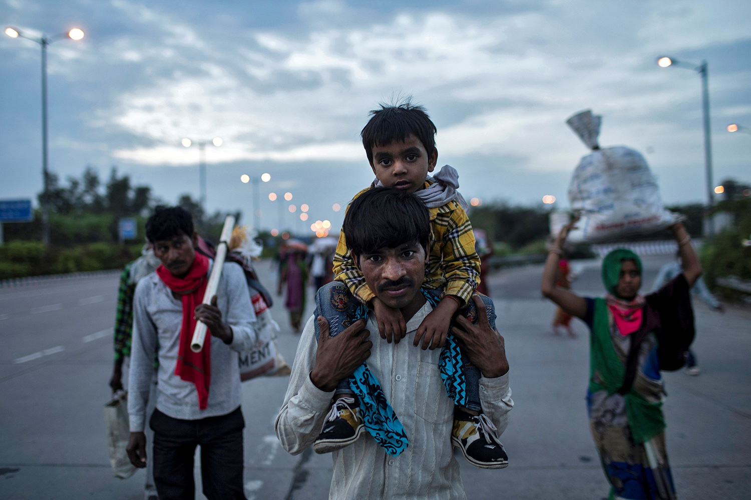 The photo shows a man with a boy on his shoulders walking with others on a road at dusk.