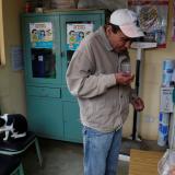 A man wearing a khaki jacket and ball cap takes medication with a glass of water.