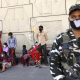 An Indian soldier in a camouflage uniform stands guard. Behind him migrants in colorful clothes sit in the shade of a high wall. 