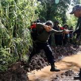 A rebel soldier helps a young female soldier cross a muddy stream surrounded by dense green jungle.