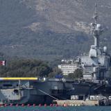 An aircraft carrier waving the French tricolor flag is docked at a port against a backdrop of mountains.