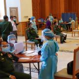 Nurses in turquoise medical gowns administer vaccines to military officers in green uniforms bedecked with medals.