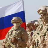 Russian soldiers in khaki-colored combat gear bearing rifles stand in a line in the desert with a Russian flag.