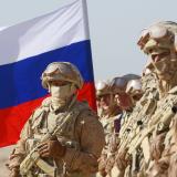 Russian soldiers in khaki-colored combat gear bearing rifles stand in a line in the desert with a Russian flag.