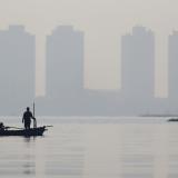 A fisherman stands on a wooden boat as smog covers high rise buildings seen in the background, at the north coast of Jakarta, Indonesia, on August 28, 2021. 