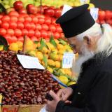 An orthodox priest with a long white beard wearing traditional black robes and hat is seen examing fresh fruits and vegetables at a market stall