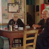 a group of four elderly greek men sit around a wooden table at a small village cafe, From left to right: the first man is wearing a white and red checked shirt with jeans, the next two are wearing black half zip shirts, the fourth man is an Orthodox priest in traditional garments