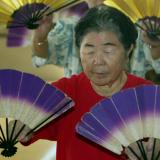 An elderly japanese woman wearing a red t shirt dances holding white and purple fans, in a group