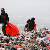 Two men wearing black jackets and pants carrying red plastic bags search a landfill for recyclable plastics