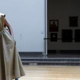 A model, wearing a khaki colored sleeveless trench coat made from recycled plastics, and blue knee high socks, strikes a pose in a dance hall as part of an art installation