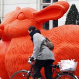 A woman on a bicycle wearing a gray jacket bikes past two giant bright orange plastic rabbits made from recycled materials
