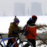 Two young boys, on in a red coat with a hockey stick and one in a brown coat with a blue bicycle walk along a snowy path next to a river. On the other side of the river, shrouded in fog are six cooling towers of a nuclear power plant