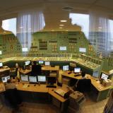 Through a large glass window, the viewer can see the silhouetted reflection of the photographer and another figure superimposed over nuclear power plant workers sitting in front of computer screens in a mint green control room the walls of which are covered in dials and knobs