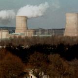 Four nuclear reactors, two dormant and two with columns of white smoke, stand against a blue sky. In the foreground are the bare branches of a forest in winter surrounding some small light-colored houses