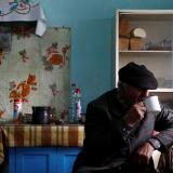 An old Belarusian man in a black hat drinks tea in his Soviet-style kitchen, which has turquoise walls, shelves with bread, and white tiles with red flowers above a small stove. It looks as though it hasn't changed since the 1980s.
