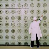 A nuclear plant worker in a white lab coat and medical hat adjust knobs on a pale green wall covered with number dials at the Windscale Pile One nuclear power plant. 