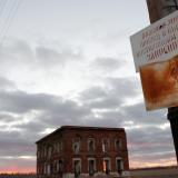 A rusted sign forbidding the gathering of mushrooms, picking berries and fishing is seen in front of an abandoned school which is silhouetted by a cloud-covered blue and orange sunset