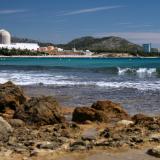 A view from a rocky beach on Spain's coastline. Across a crescent of turquoise ocean, waves cresting with white foam, can be seen the Vandellos II nuclear power plant sitting between green hills