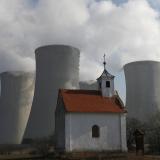 A small white chapel with a red roof stands in front of four concrete reactor towers, each billowing white smoke. 