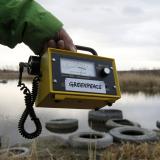 An extended arm in a green coat sleeve holds out a yellow Geiger counter, which shows a high level of radiation over the the banks of the Techa river, which is relatively still. Its banks are covered with old rubber tires and tall yellow grass