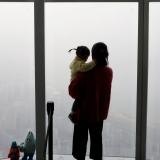 A woman holding her baby in her arms looks out a window at the city shrouded by a fine dust during a polluted day. 