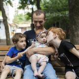 A young adult man is pictured with his three small children on a park bench.