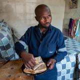 A middle-aged man is pictured eating a ham and cheese sandwich on the corner of his bed. 