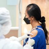 A woman wearing a blue shirt receives a COVID vaccine from a medical provider in a white gown.