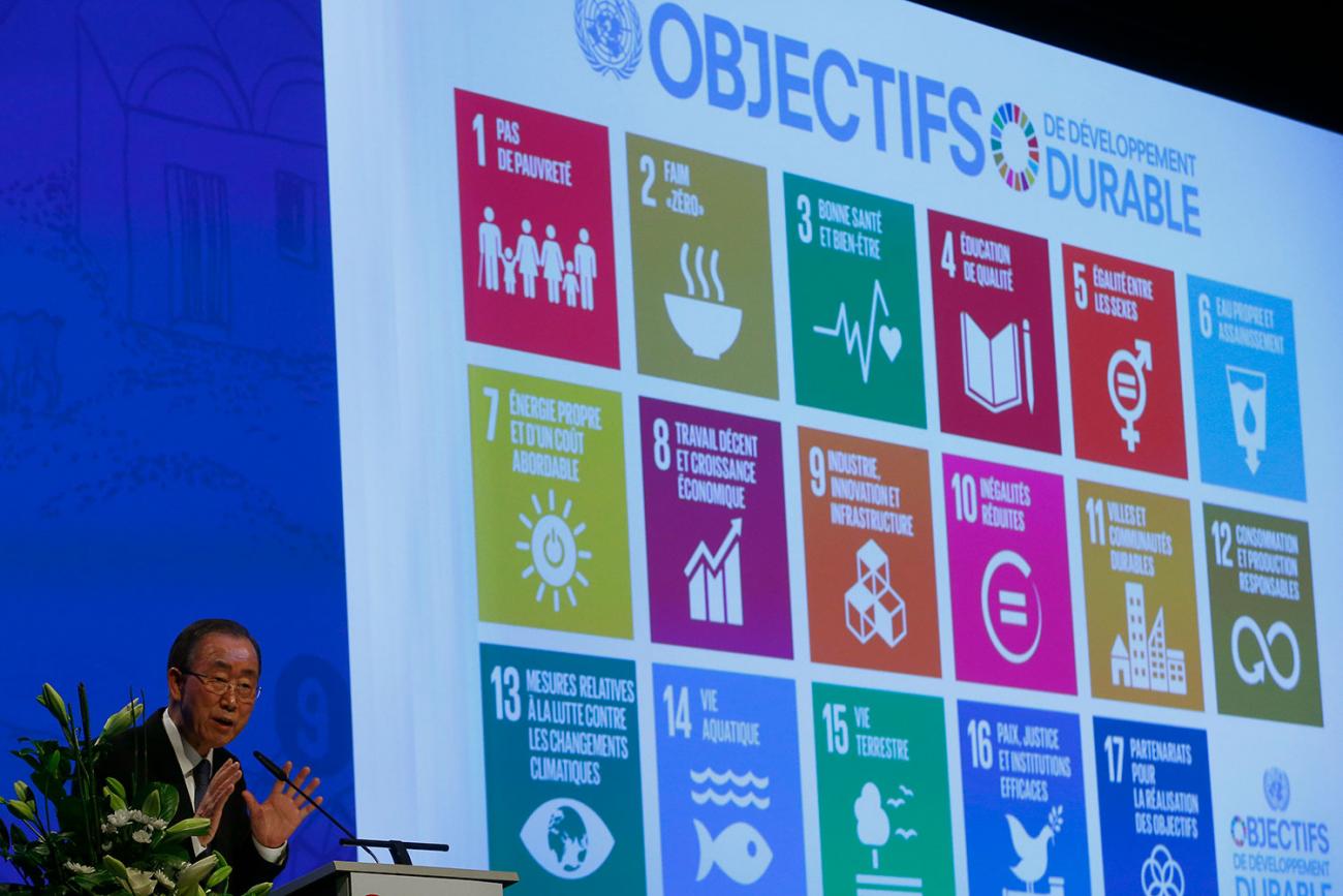 United Nations Secretary-General Ban Ki-Moon sits at a table on a stage with a large screen. The photo shows him in the corner of the frame. On the screen behind him are displayed pastel-colored icons representing the 17 goals of UN's 2030 Agenda for Sustainable Development.