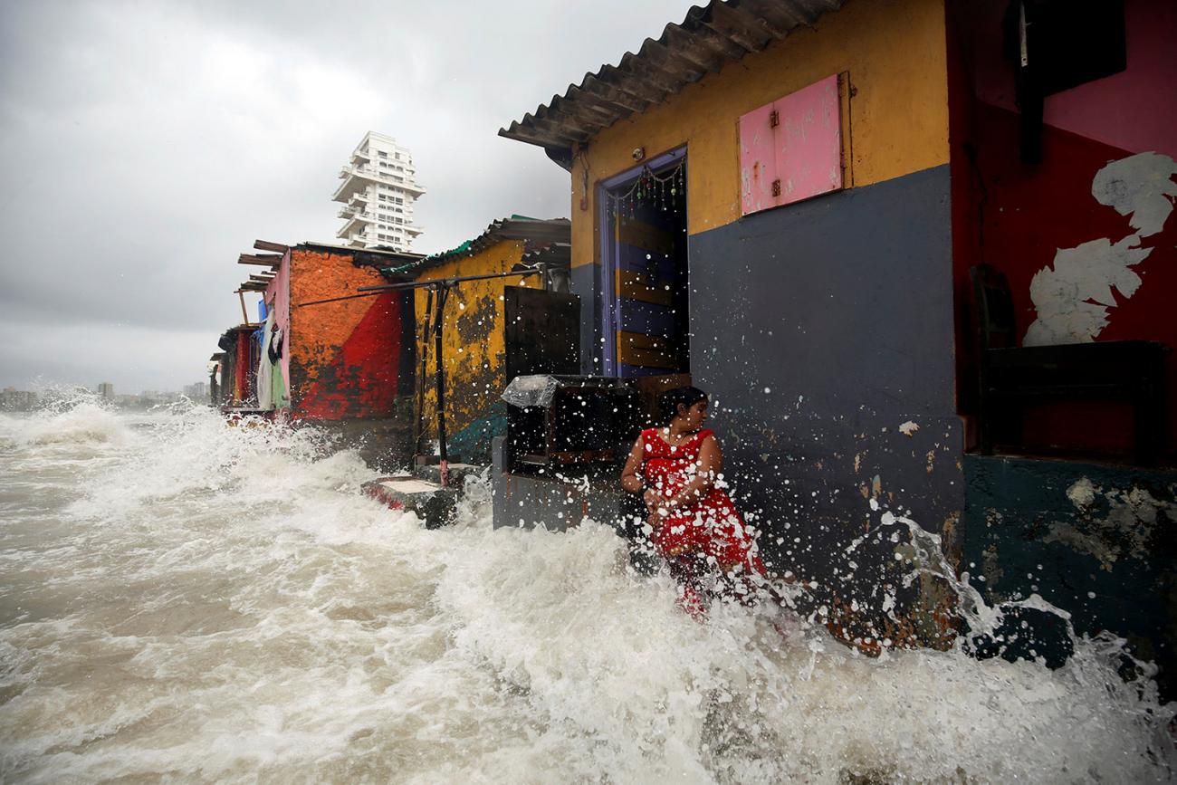 A woman is standing in front of a red, yellow, and grey slate colored tin-roof building with an open door. It is one of several structures in a row—there’s an orange one, a green one, and a yellow one. She is turning her head away from the violence of the crashing wave, which sends water shooting up all around her and nearly to the top of the building. All the area in front of the structure is covered by turbulent water. This is a frightening scene.