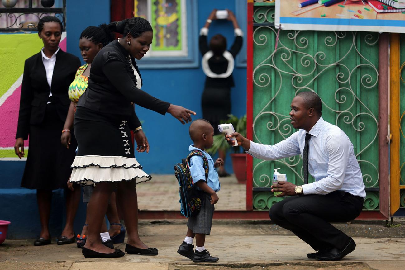 Picture shows a young boy carrying a backpack and wearing a school uniform standing in front of a brightly-colored building. A well-dressed woman directs him while a well-dressed man kneels and points a high-tech thermometer at his head to take his temperature.